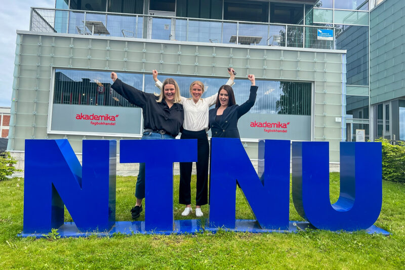 Tre studenter står bak NTNU-bokstavene på campus, smilende, med hendene i været. foto