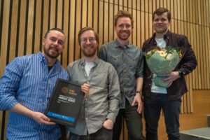 Bildet viser fire av de seks ansatte i Ambolt Audio. De holder opp prisen og blomsterbuketten.