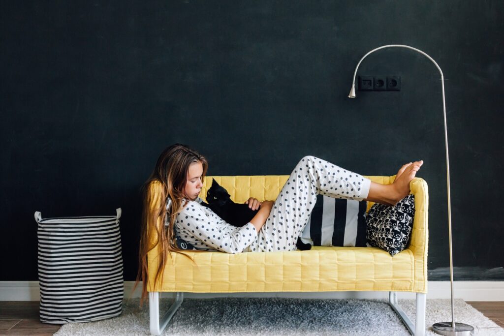 Portrait of child girl wearing pajama hugging her cat on the yellow couch against black wall in modern living room interior