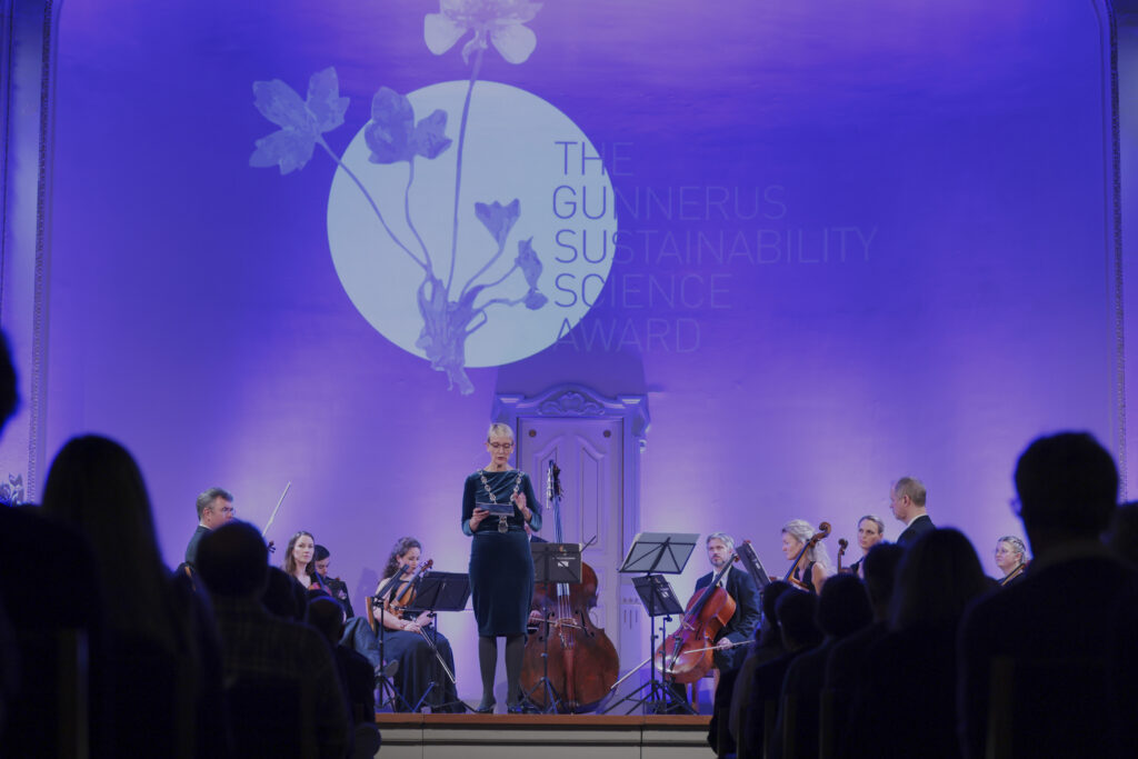 Rektor Anne Borg er festkledt, står på en scene foran et orkester, og taler mot et publikum