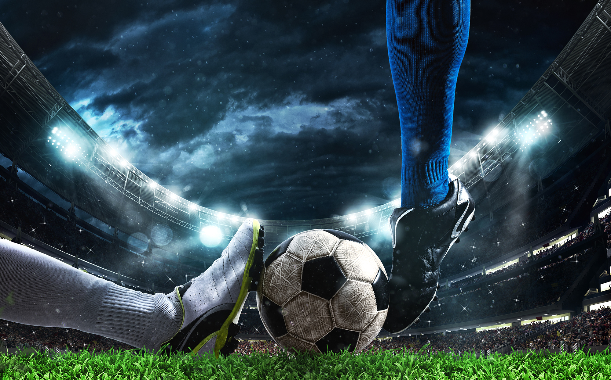Illustrasjon. Nærbilde av en fotball som ligger på en gressmatte, to ben, med forskjellige fotballsko prøver fra hver sin kant å bevege ballen. I bakgrunnen ser man tribunene på fotballstadion. himmelen er mørk