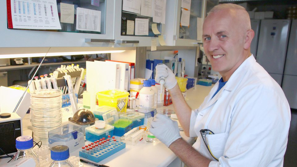 Man in white coat sitting by a laboratory bench.