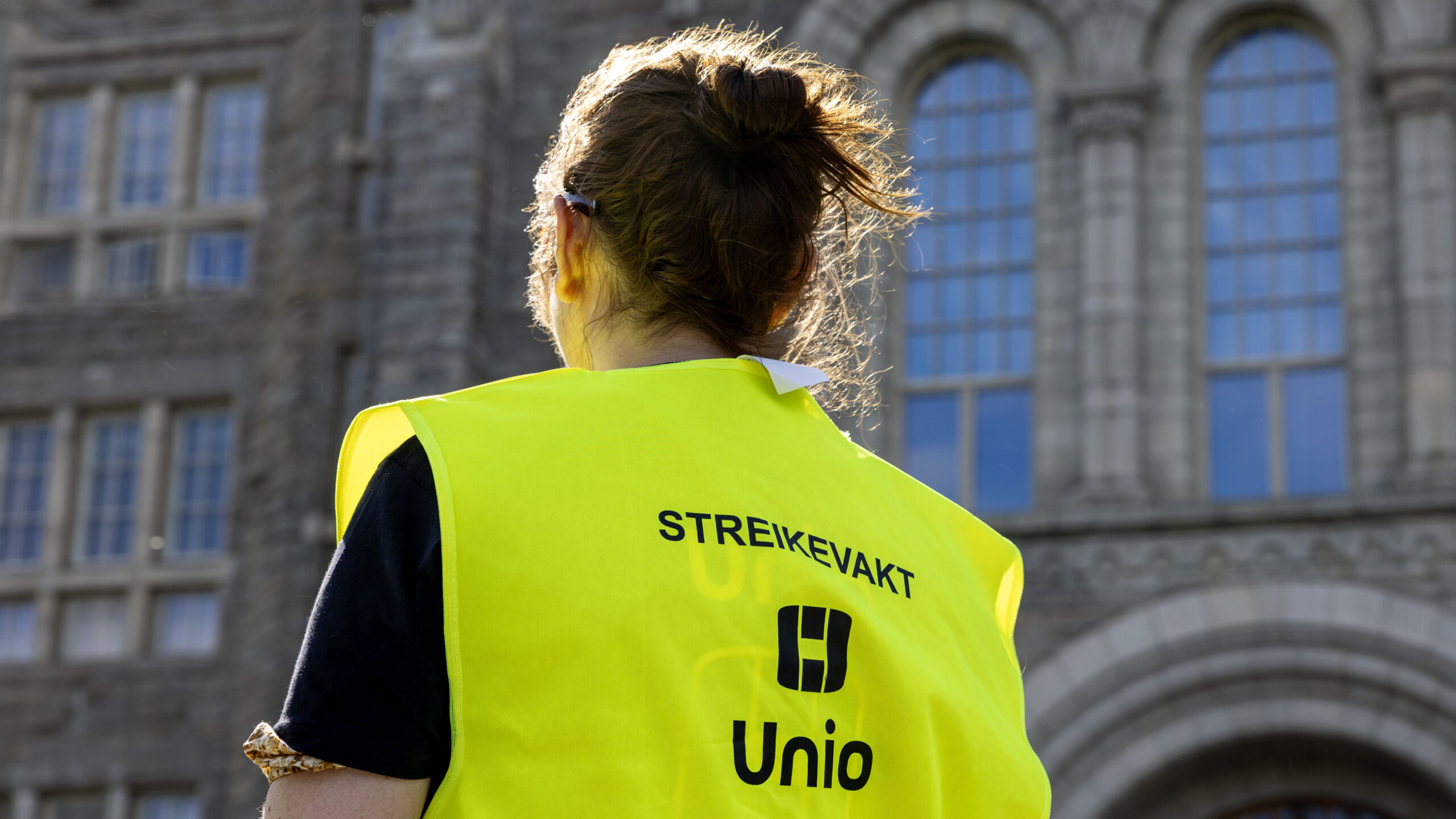 En person står med ryggen til kamera. Hun har på en gul vest der det står "Streikevakt Unio"