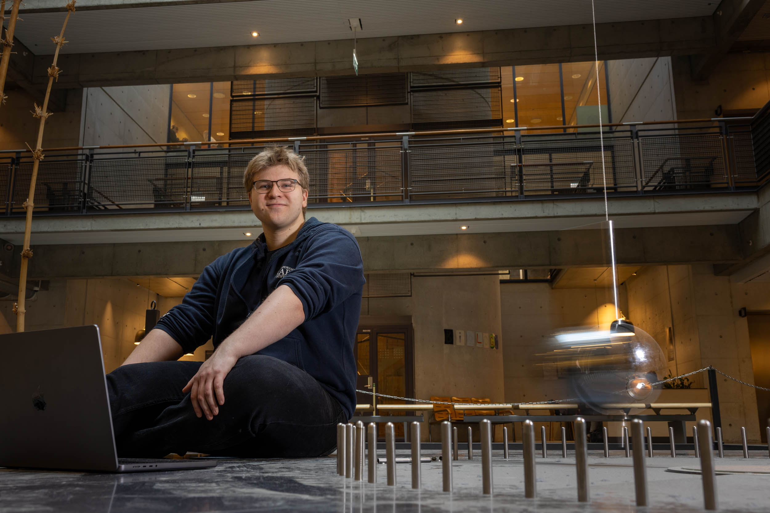 Ung mann sitter ved en pendel i bevegelse i Realfagbygget på NTNU-campus på Gløshaugen i Trondheim.